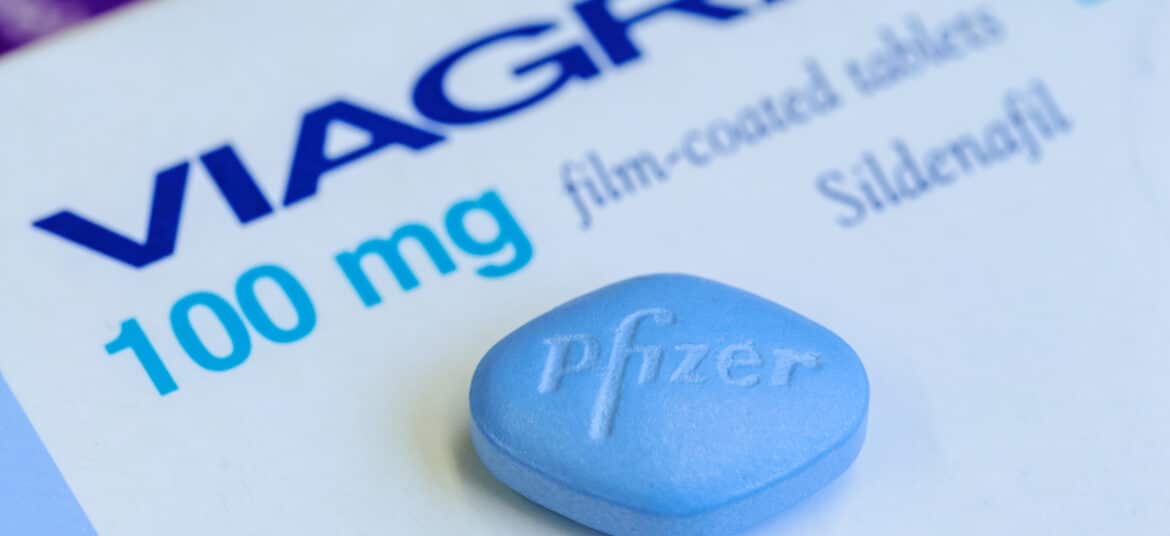 Imagem editorial de um comprimido azul de Viagra, produzido pela Pfizer, posicionado ao lado de sua embalagem original, com o logotipo da Pfizer visível.