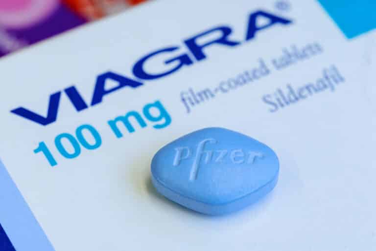 Viagra: Tudo que você precisa saber 13 Imagem editorial de um comprimido azul de Viagra, produzido pela Pfizer, posicionado ao lado de sua embalagem original, com o logotipo da Pfizer visível.