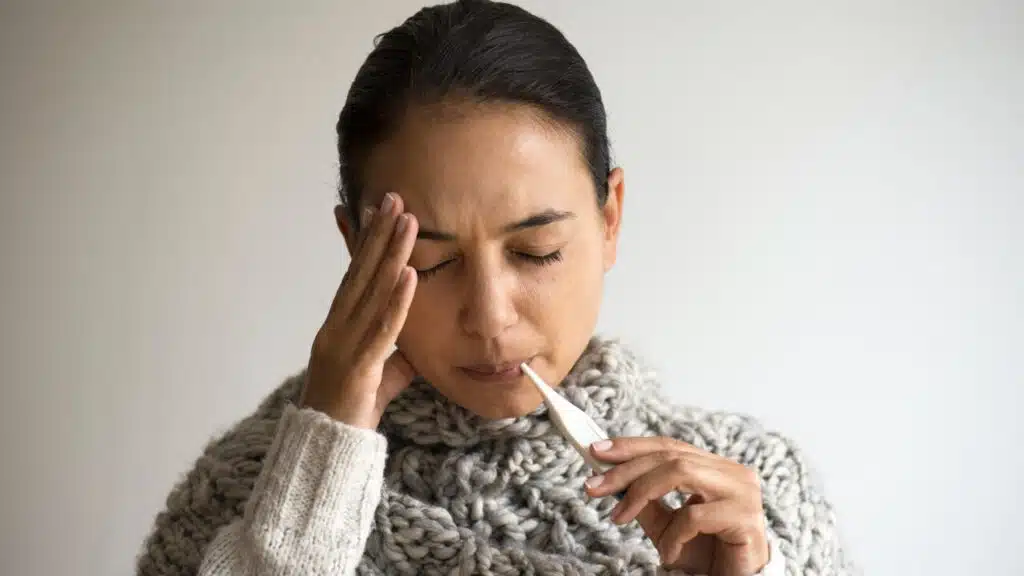 mulher com a mão na cabeça com dor e um termômetro na boca para medir sua febre maculosa adiquirida por uma bactéria