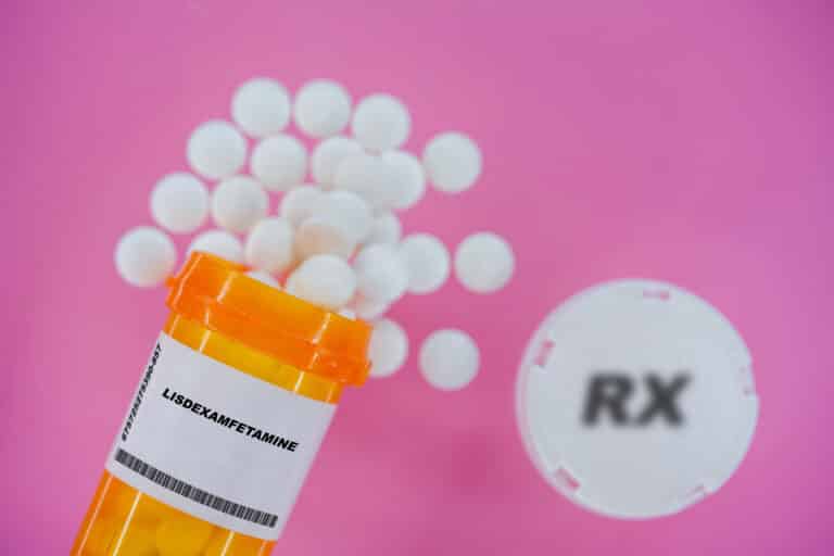Venvanse: O Guia Completo para Entender Este Medicamento 3 Comprimidos de medicamento prescrito Lisdexamfetamina em um frasco de plástico com comprimidos. Pílulas derramando de um recipiente amarelo sobre um fundo rosa.