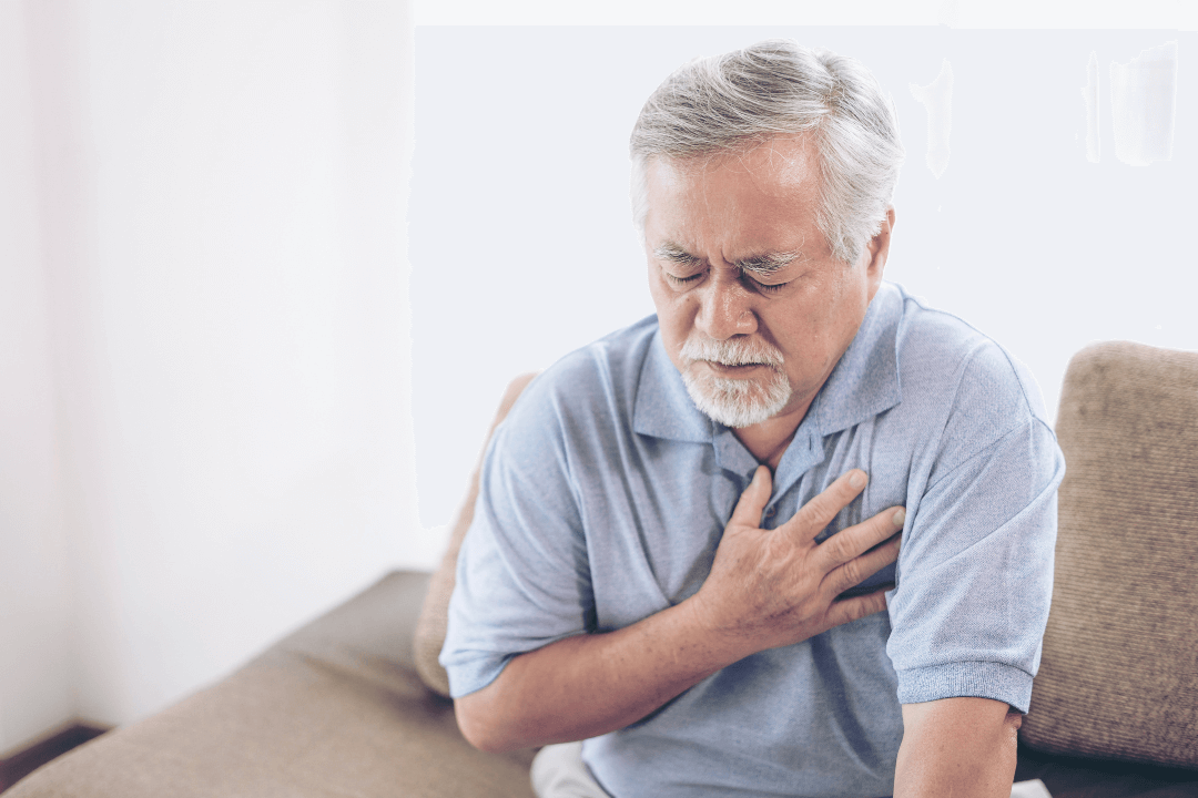 um senhor está sentado no sofá com as mãos no peito, principiando um infarto