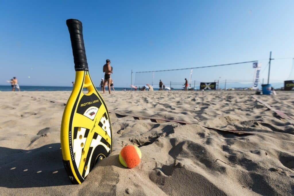 raquete de beach tennis e uma bola colocadas na areia da praia e pessoas jogando ao fundo
