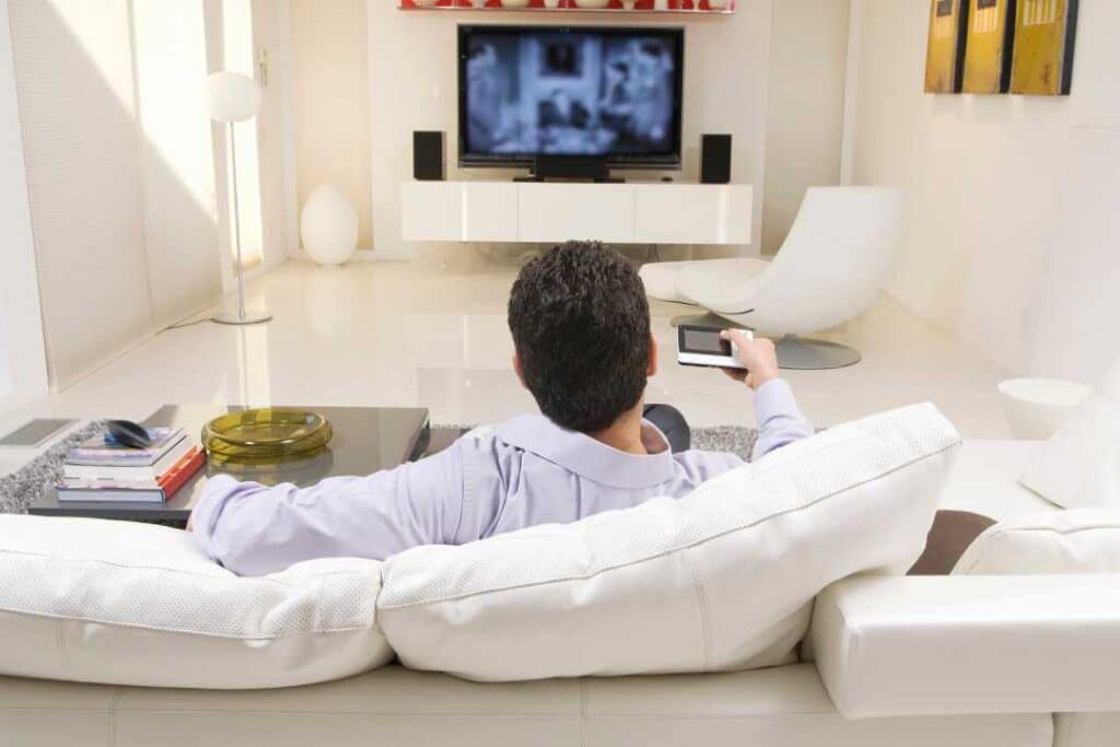 homem sentando em um sofá em uma sala e apontando um controle para uma televisão